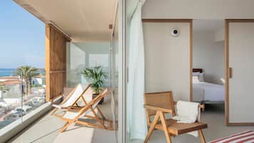 A balcony with a view of the sea, two wooden lounge chairs, a potted plant, and a bed with white bedding.
