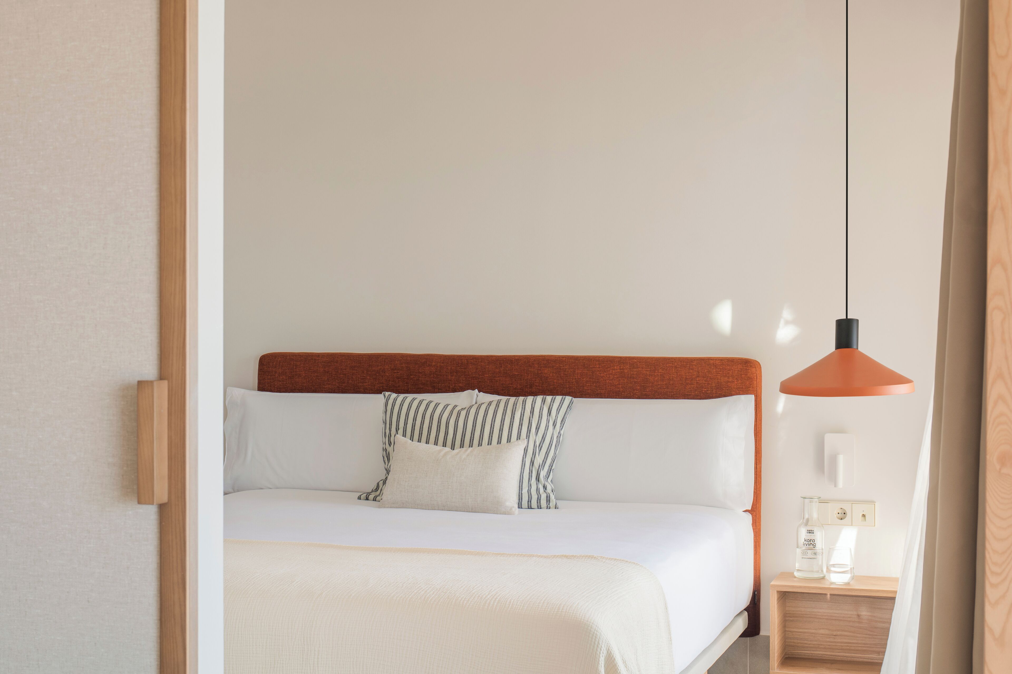 A neatly made bed with white bedding and striped pillows, a wooden headboard, and a bedside table with a bottle and a glass.