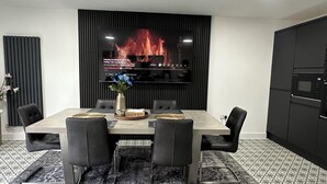 A modern dining room with a large TV displaying a fireplace scene, a vase with flowers, and a patterned rug. - Luxurious New House Near Manchester Airport (Wythenshawe)