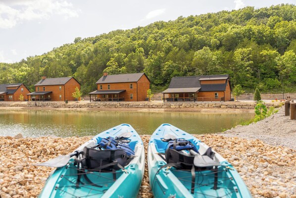 Vista de un lago con cabañas de madera y kayaks sobre piedras.
