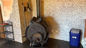A wood stove with a chimney, a broom, and a trash bin in a room with a tiled floor and a stone-patterned wall.