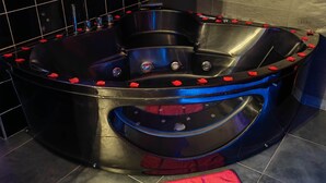 Une salle de bain moderne avec un jacuzzi noir entouré de pétales de roses rouges, un tapis de bain rouge et un mur recouvert de carreaux noirs.