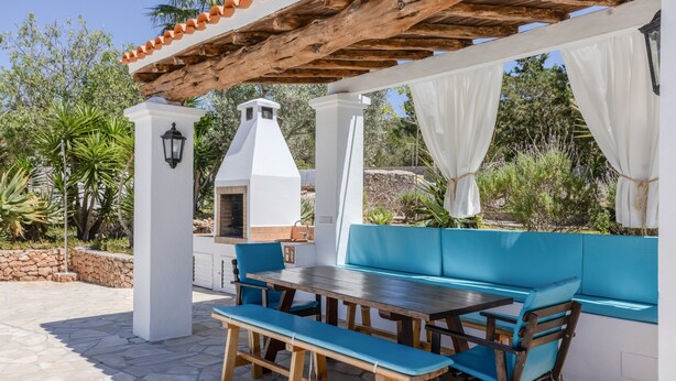 Une terrasse extérieure avec une table en bois, des sièges rembourrés bleus, un sol en pierre et un barbecue blanc.