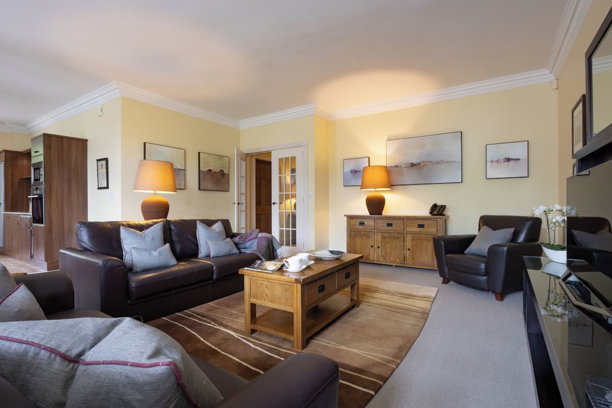 A living room with a sofa, armchairs, a coffee table, and a TV. There are lamps, framed pictures, and a cabinet.