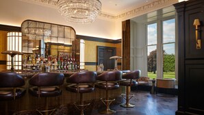 A bar area with multiple stools, a chandelier, and a bar stocked with bottles. - Mansion House Apartments at Cameron Lodges (Alexandria)