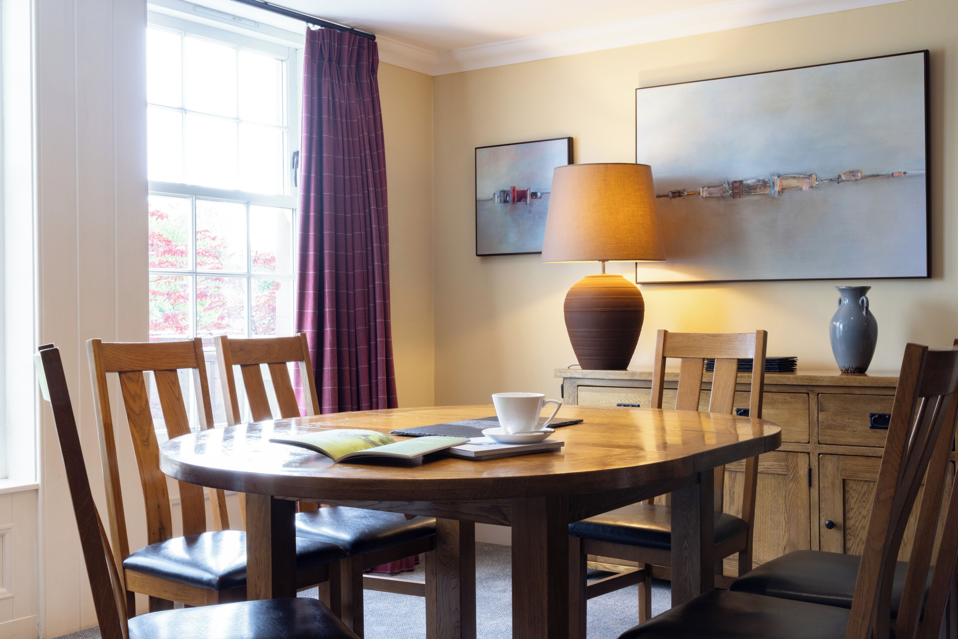 A dining area with a round table, chairs, a lamp, a vase, and artwork on the wall.