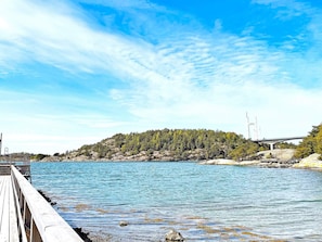 Une jetée en bois s’étendant dans une eau calme, une côte rocheuse bordée d’arbres et un pont visible au loin.