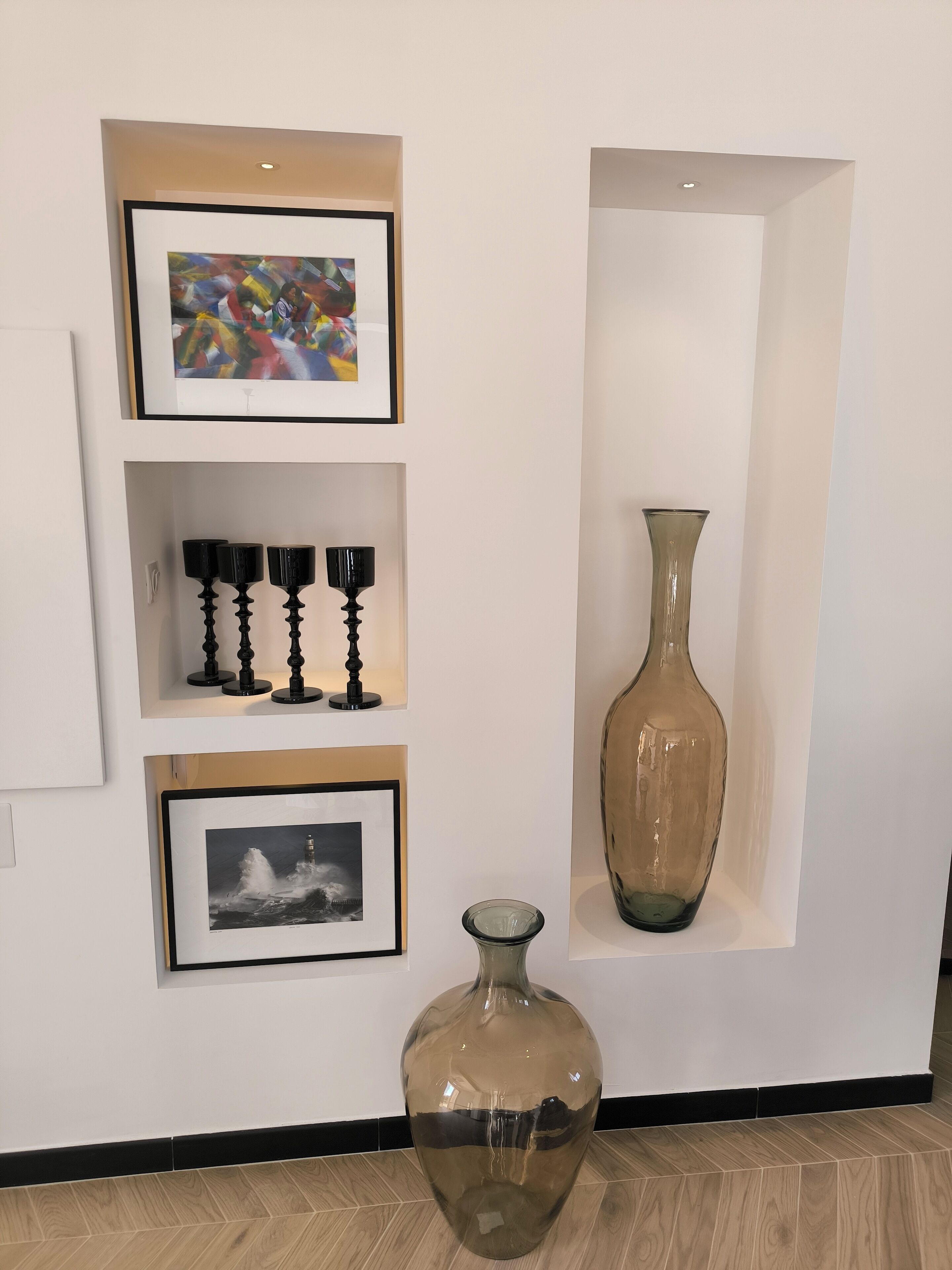 Een muur met drie ingelijste kunstwerken, een hoge vaas en een grote vaas op de vloer.