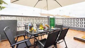 Un patio exterior con una mesa lista para dos personas, rodeado de sillas y cubierto por una sombrilla grande.