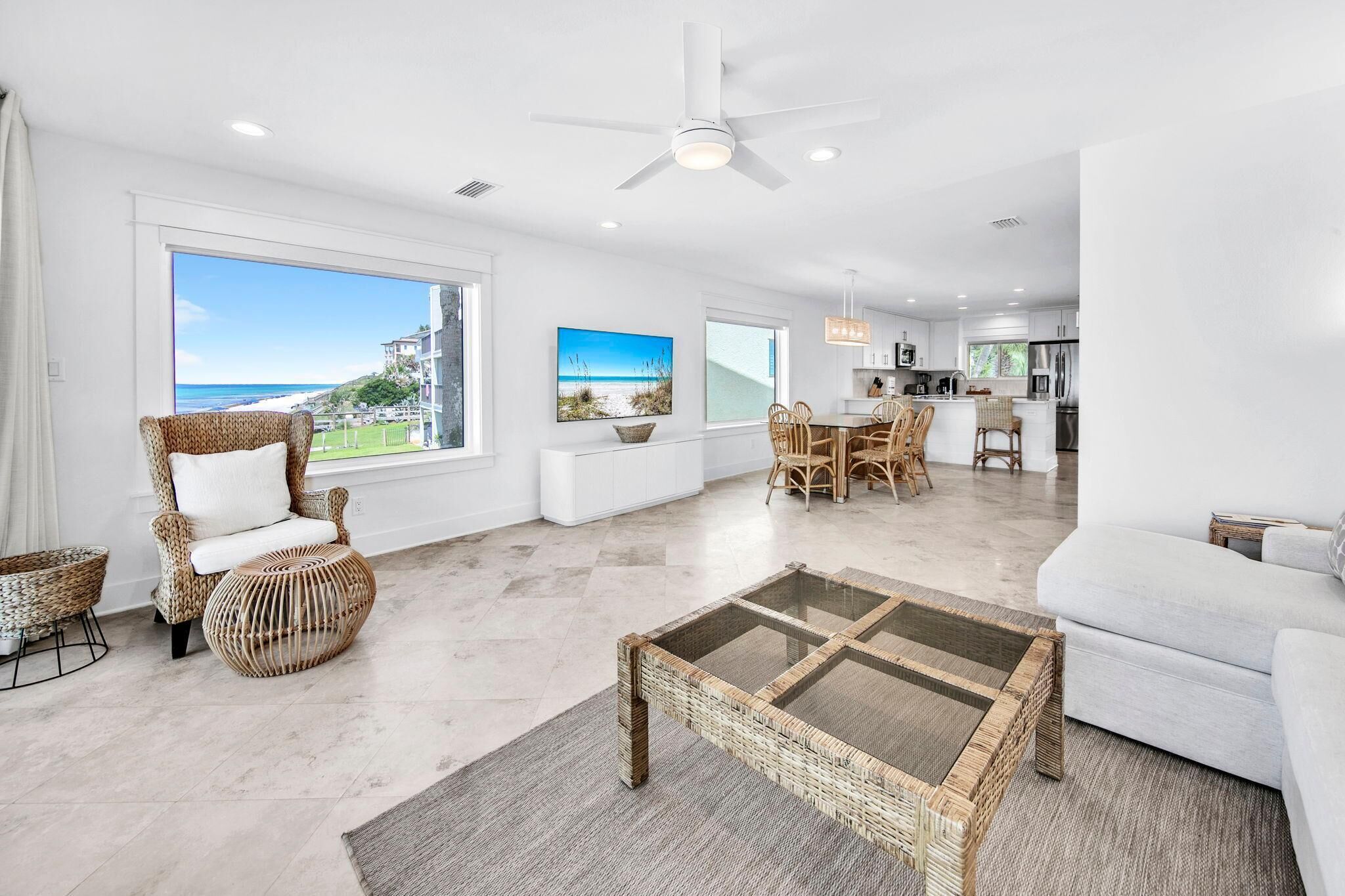 A spacious living area with a large window overlooking a beach, wicker furniture, a glass-top coffee table, and a modern kitchen in the background.