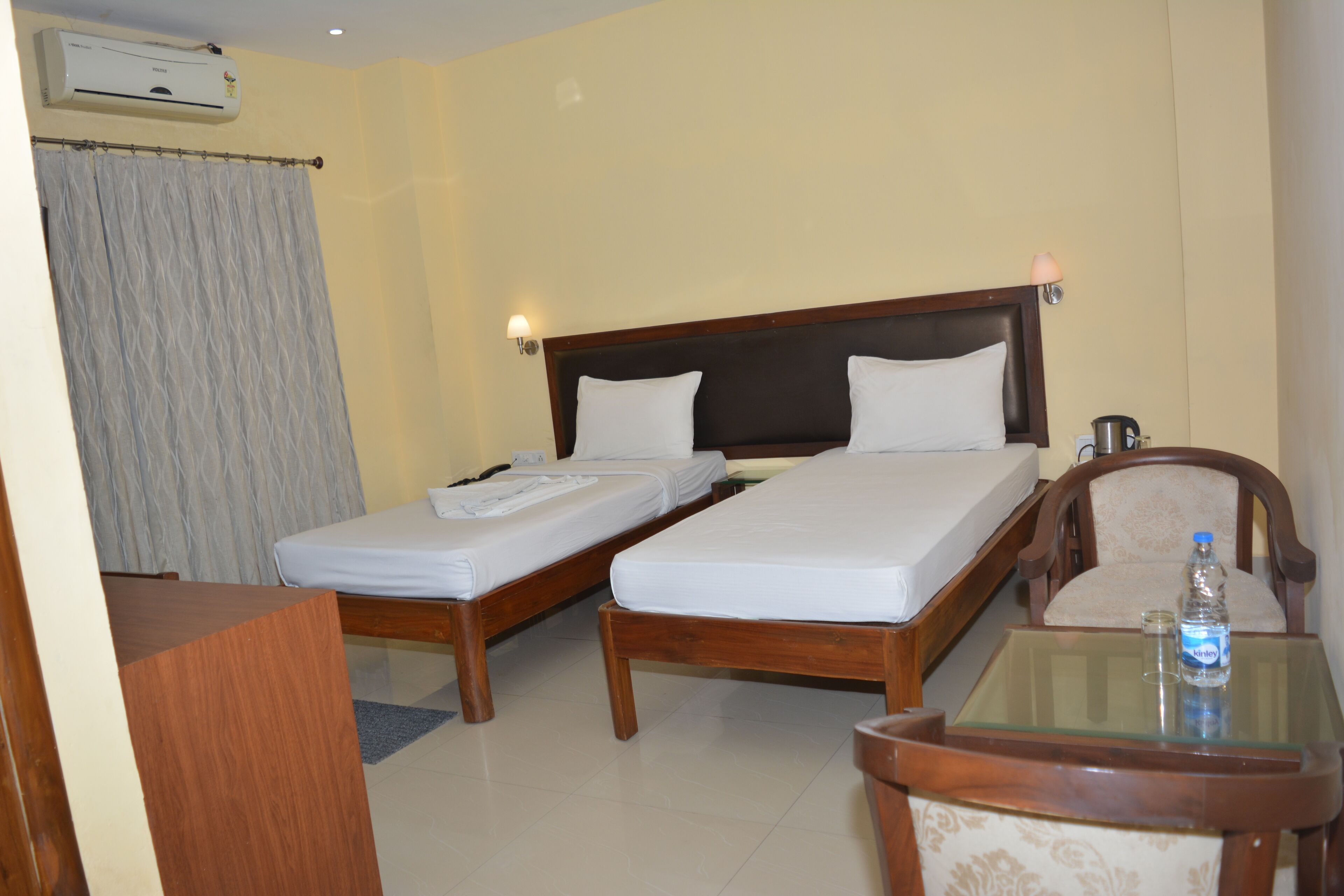 A hotel room with two single beds, a wooden nightstand, a small table with a bottle, and a chair.