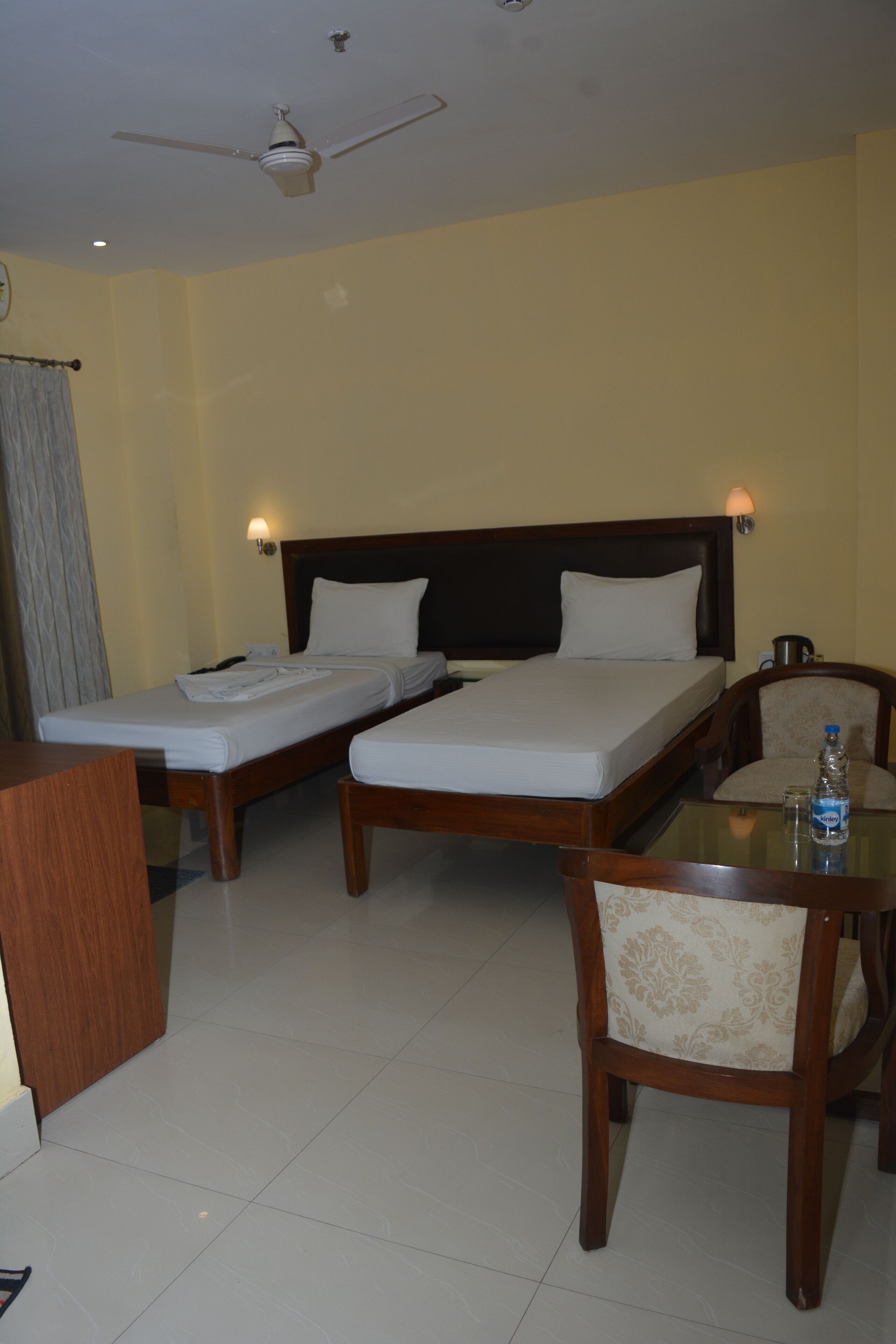 A hotel room with two single beds, a wooden nightstand, a table with a bottle, and a ceiling fan.