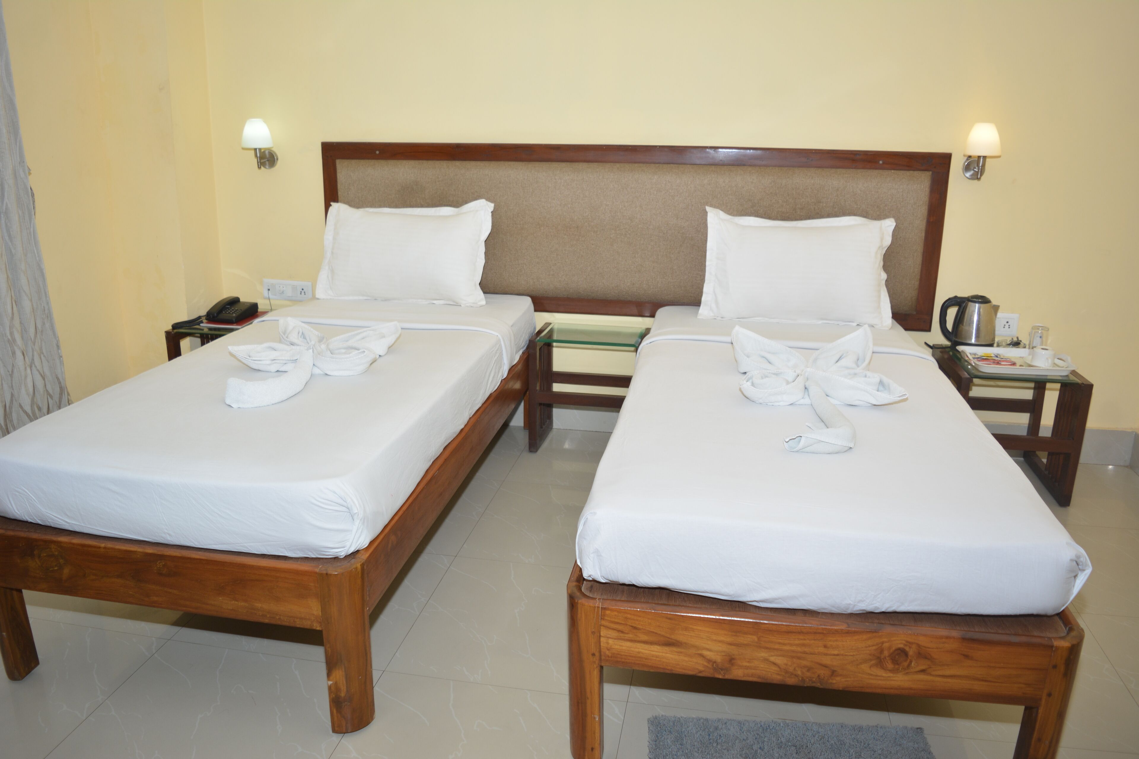 A hotel room with two single beds, wooden headboards, white linens, a bedside table with a lamp, a telephone, and a coffee maker.