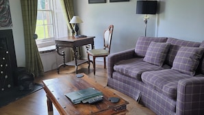 A living room with a wooden coffee table, a purple sofa, a wooden chair, a small table with a lamp, and a window with curtains.