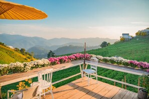 A wooden deck with white chairs and a table, overlooking a lush green landscape with a row of colorful flowers and distant mountains. - Phuwin Homestay Monjam (Mae Rim)