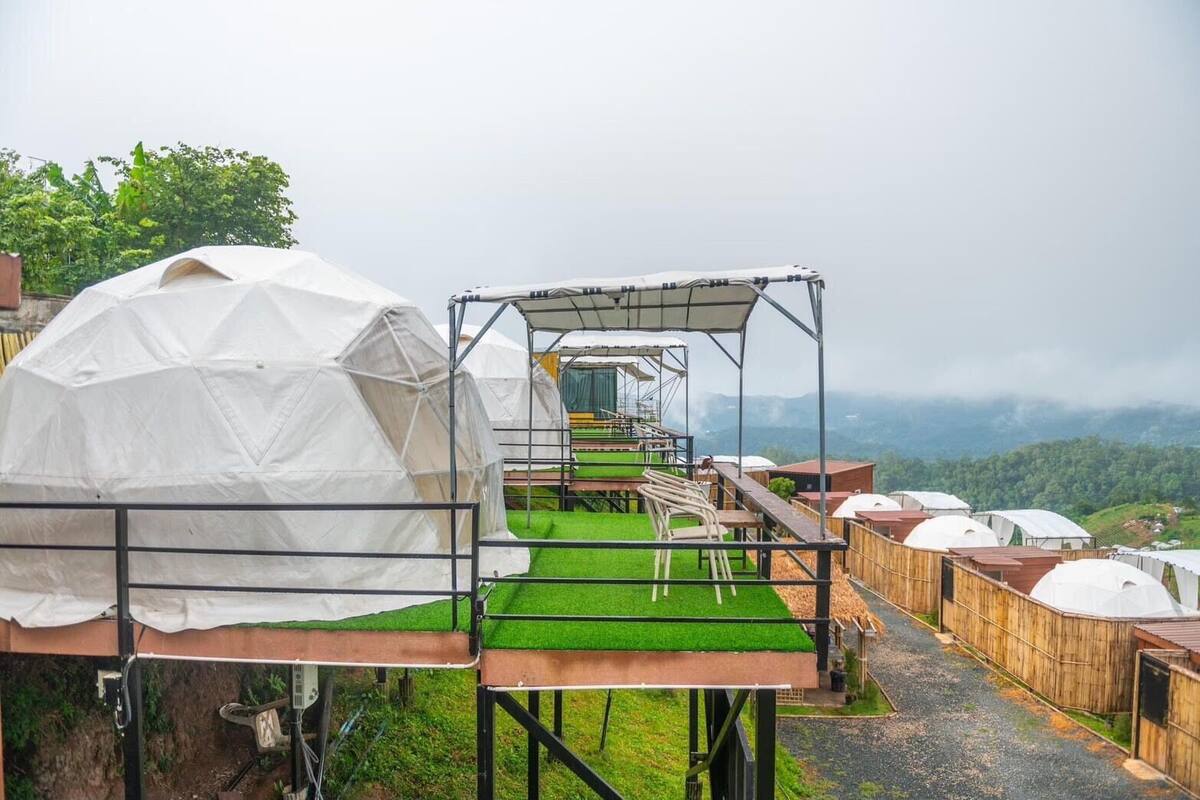 A series of dome-shaped tents with a covered walkway connecting them, set against a backdrop of green hills.