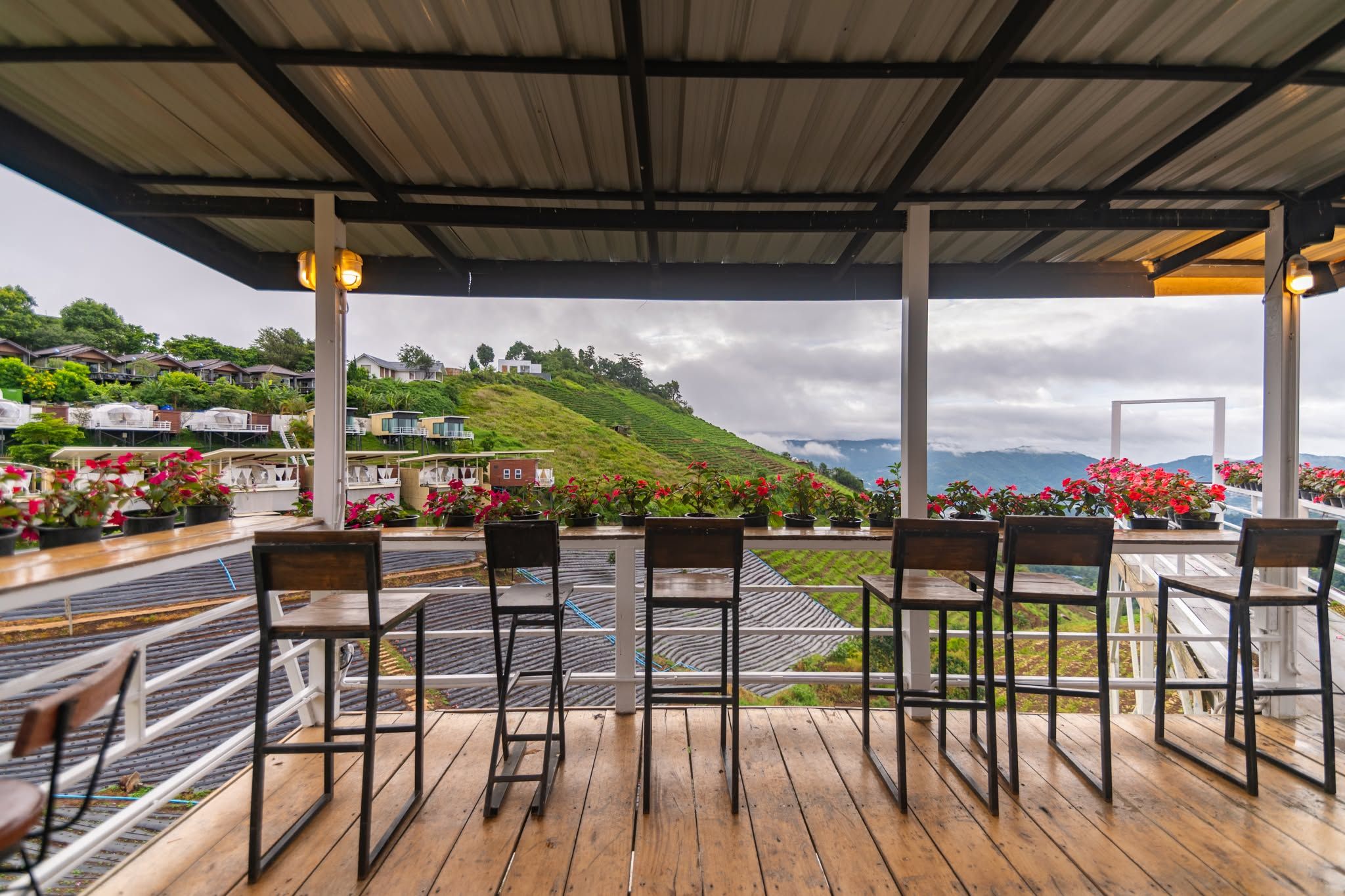 A covered outdoor seating area with chairs and tables, overlooking a hillside with houses and a railway track.