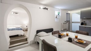 A modern hotel room with a bed, a dining table with breakfast, a kitchen area, and a bathroom.