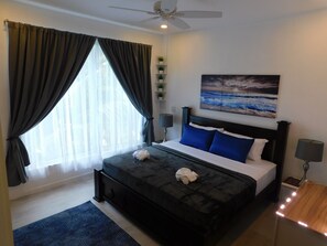 A bedroom with a bed, dark curtains, a ceiling fan, a painting, bedside lamps, and a small shelf with plants.