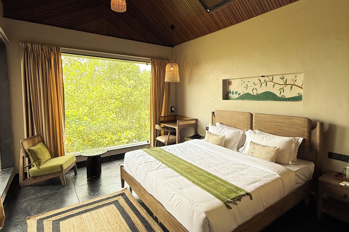 A bedroom with a large bed, a ceiling fan, a desk, and a view of greenery through the window.