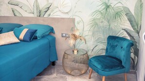 A room with two beds, a teal chair, and a round side table with a plant.