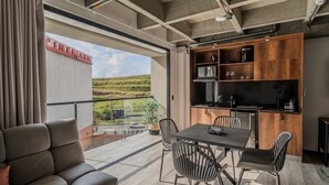 Una cocina moderna con armarios de madera, un comedor con mesa y sillas, y un balcón con vistas a un campo y un cartel de cine.