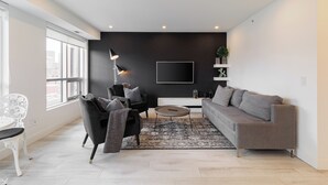 A modern living room with a grey sofa, a black armchair, a white coffee table, and a large window providing natural light. - Luxury Downtown Suite w/ Balcony Views, Air Conditioning + Free Parking (Calgary)