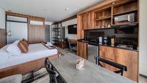 A modern hotel room with a bed, a desk, a kitchenette, and a TV.