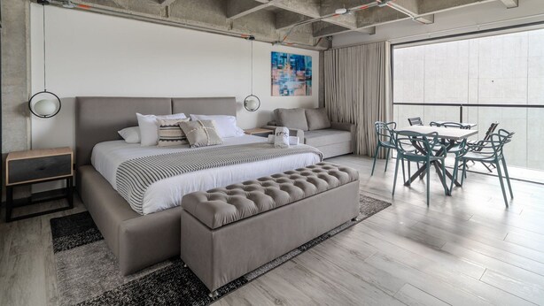 Un dormitorio moderno con una cama grande, una banca, una mesa de comedor con sillas y un ventanal amplio.