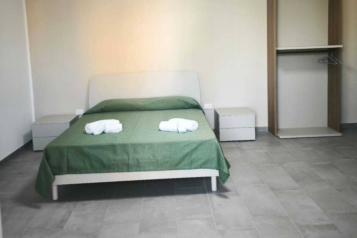 A single bed with a green bedspread, two bedside tables, a wardrobe, and a tiled floor.