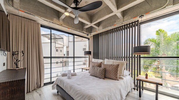 A modern bedroom with a large bed, a desk, and a balcony with a view of buildings and palm trees.