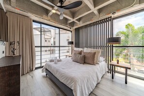 Ein modernes Schlafzimmer mit einem großen Bett, einem Schreibtisch und einem Balkon mit Blick auf Gebäude und Palmen.