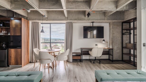 Una habitación moderna con comedor, un escritorio y una silla, un televisor montado en la pared y un amplio ventanal con vistas a la ciudad.