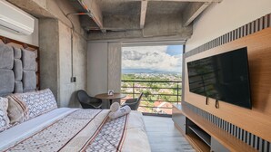 A modern hotel room with a large bed, a flat-screen TV, a balcony with a city view, and a small table with a chair.