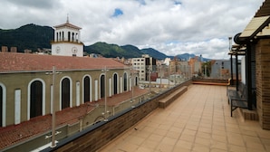 Une terrasse sur le toit offrant une vue sur un bâtiment historique doté d’arcs en plein cintre et d’un clocher, le tout entouré d’immeubles modernes et de montagnes.