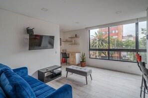 Een moderne woonkamer met een blauwe bank, een grijze poef, een aan de muur bevestigde televisie, een bureau met stoel en een groot raam met uitzicht op gebouwen en bomen.