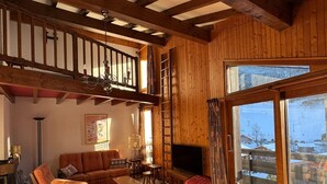 A cozy living room with a wooden ceiling, a sofa, a TV, and a large window with a snowy view.