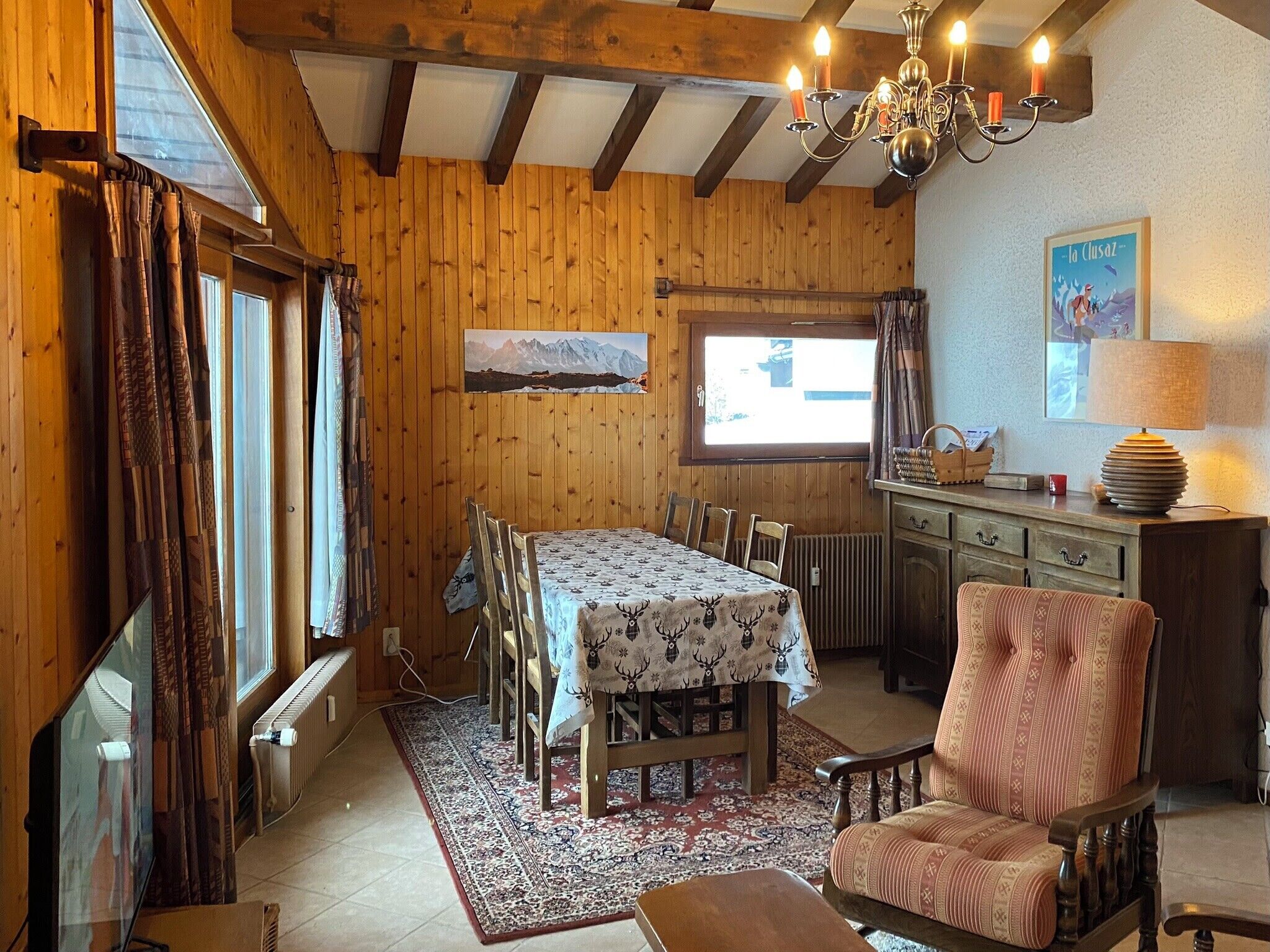 A cozy wooden cabin interior with a dining table set for four, a patterned rug, a wooden chair, and a chandelier.