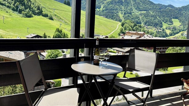 A balcony with a table and chairs, overlooking a green valley with houses and trees.