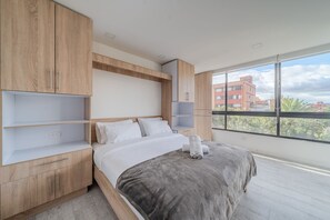 A modern bedroom with a large bed, wooden cabinetry, and a view of buildings through large windows.