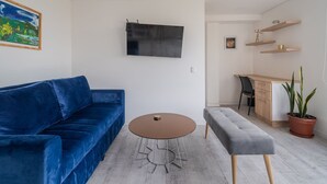 Un soggiorno con un divano blu, un tavolino rotondo, una panca grigia, una TV a schermo piatto, un mobiletto in legno e una pianta in vaso.