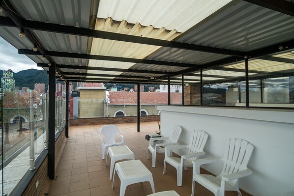 A covered balcony with white plastic chairs and tables, overlooking a cityscape with buildings and a mountain.