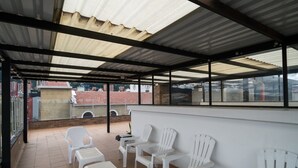 Uma área de estar coberta ao ar livre, com cadeiras e mesas de plástico branco, telhado de metal e vista para edifícios ao fundo.