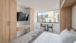 Quarto moderno com cama grande, TV fixada num armário de madeira, mesinha com cadeiras e vista para edifícios pela janela.