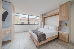 Et moderne soveværelse med seng, træskab og stort vindue med udsigt til bygninger.