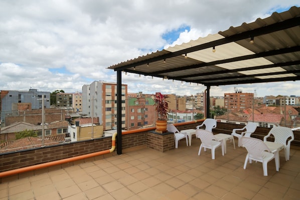 A rooftop terrace with a covered seating area, potted plants, and a view of surrounding buildings.