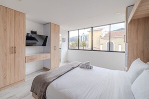 A modern bedroom with a large bed, a TV on a wooden cabinet, and a city view through a large window.