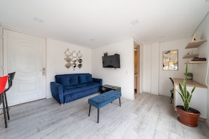 Een moderne woonkamer met een blauwe bank, een klein bijzettafeltje, een aan de muur bevestigde televisie en een potplant.