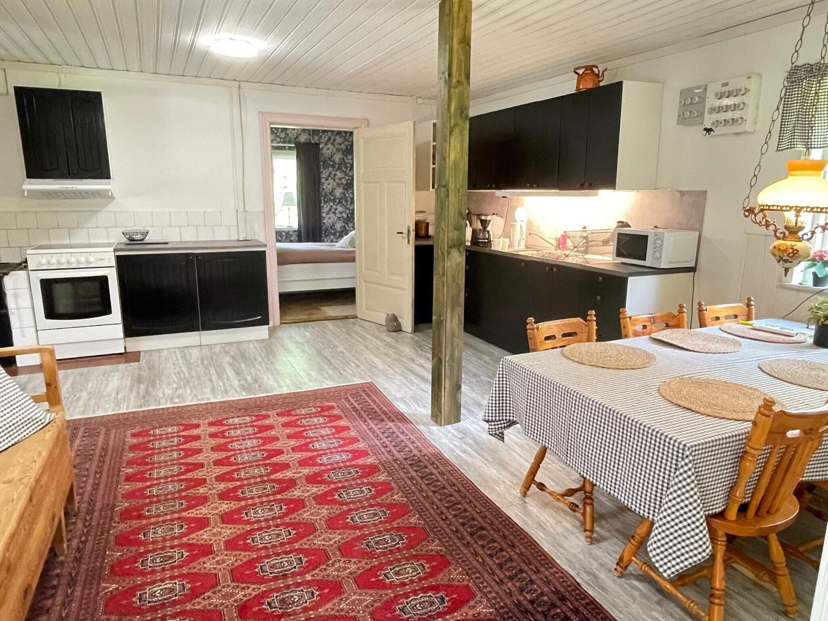 Een compacte keuken met een fornuis, magnetron en zwarte kastjes. Een zithoek met een geruit tafelkleed en stoelen is zichtbaar.