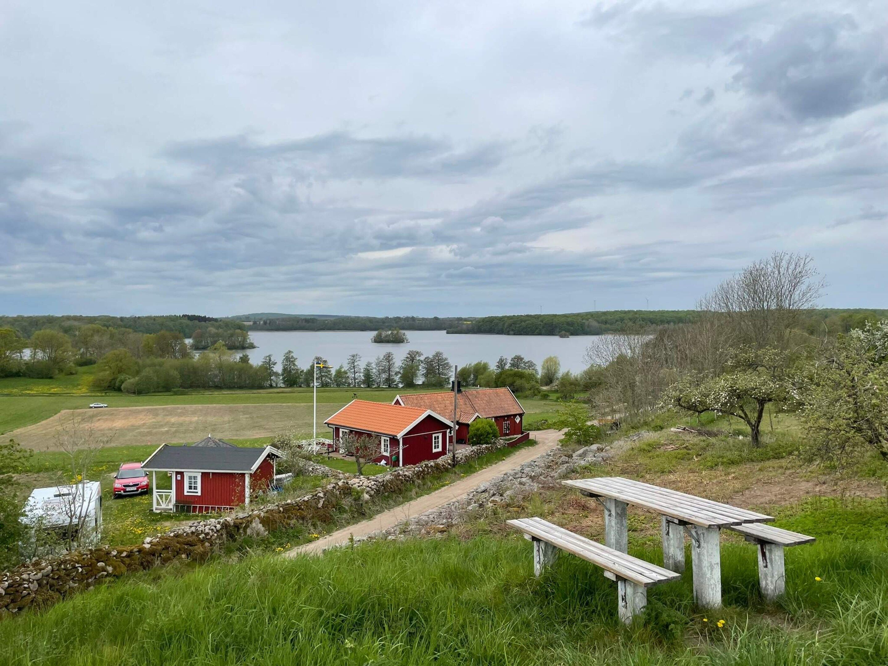 A rural landscape with a lake, a red house, a white car, and a picnic table.